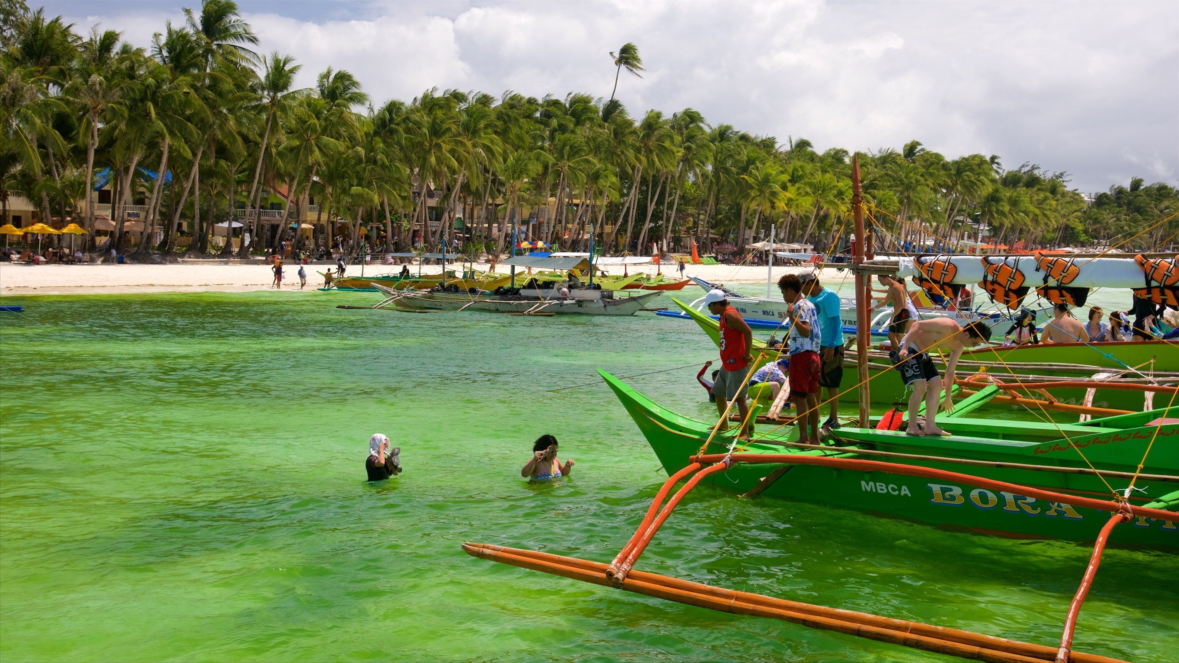 A tropical beach with clear green water, palm trees, and traditional boats.