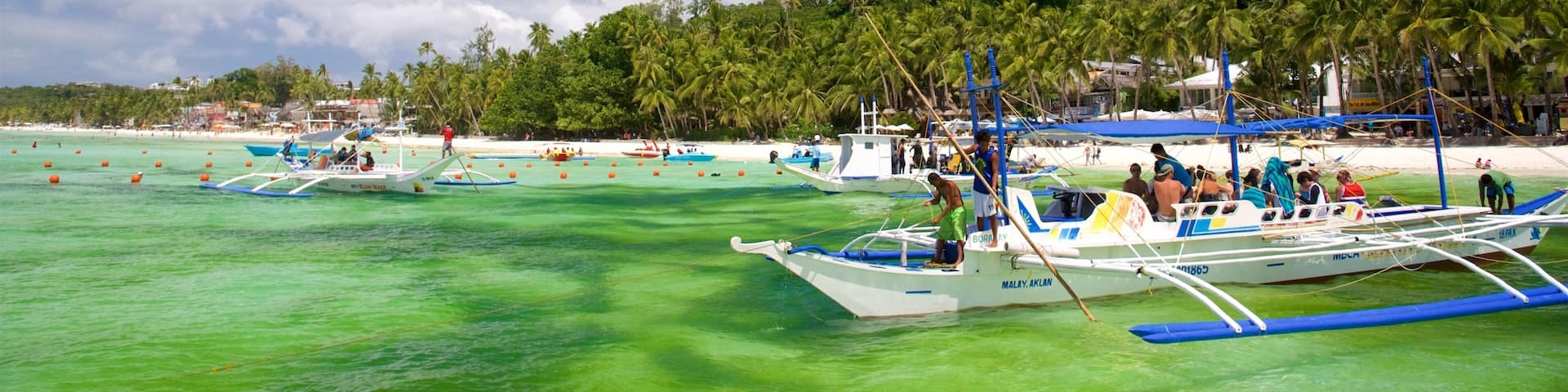 A beach with clear turquoise water, several boats, and a hill with palm trees in the background.