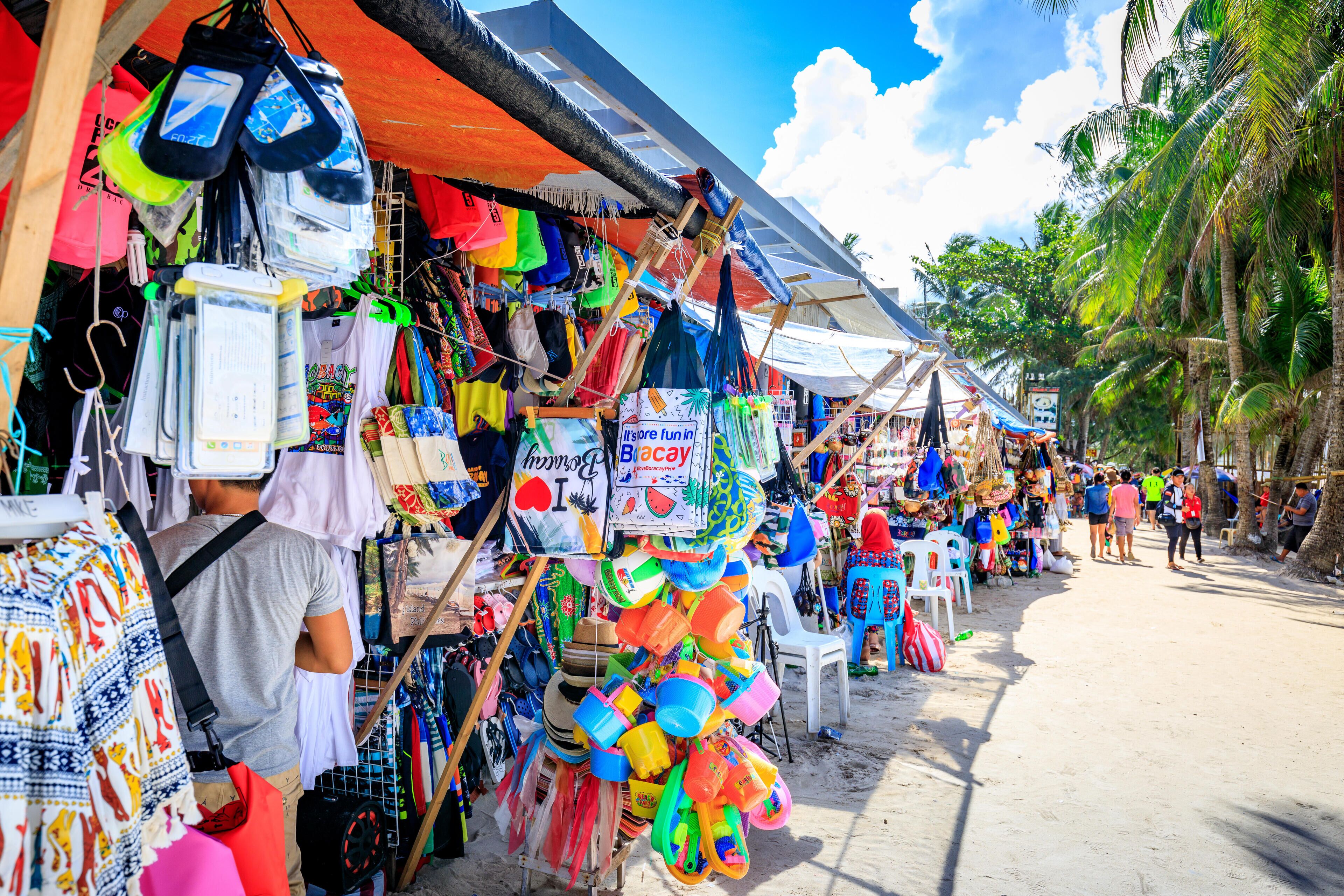 A beachside market with colorful beach gear and a sign that reads "It's fun in Bacay."