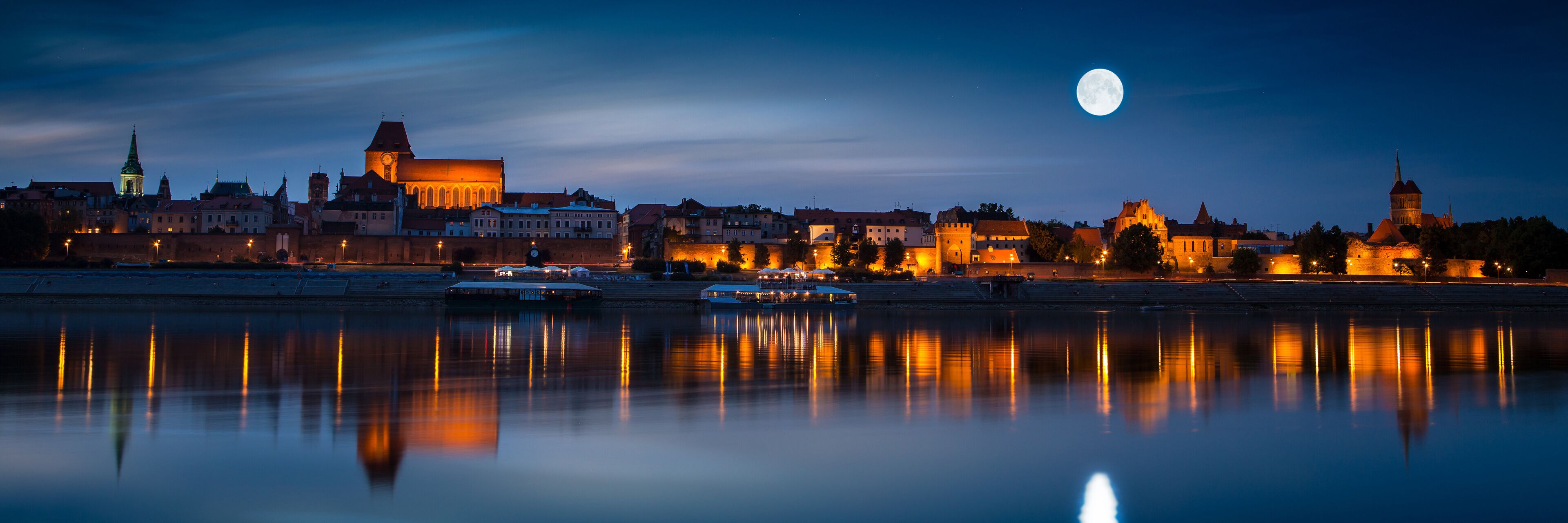 A cityscape at night with a calm river reflecting lights, a prominent church, and a full moon in the sky.