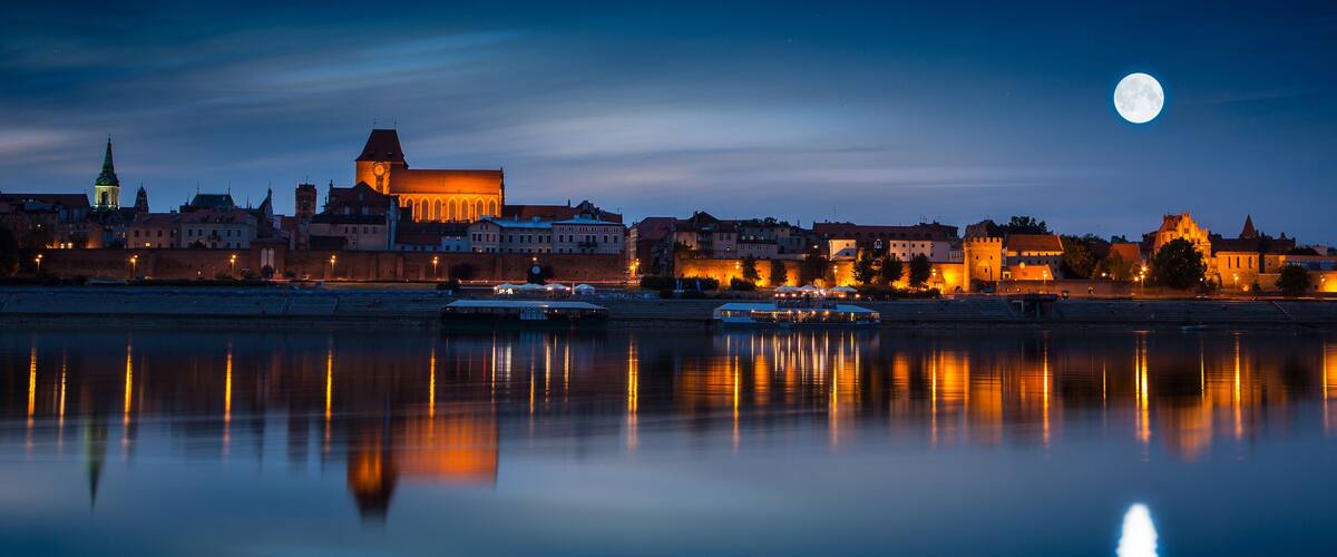 A cityscape at night with a calm river reflecting lights, a prominent church, and a full moon in the sky.