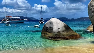 A clear turquoise sea with boats, a large rock in the foreground, and mountains in the background.