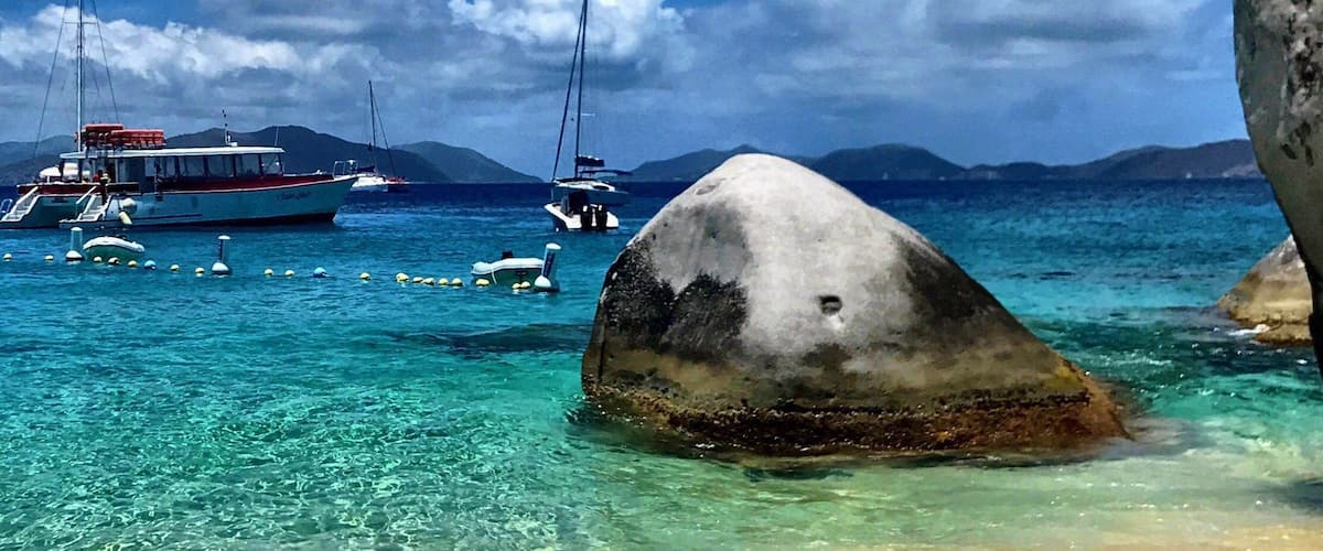 A clear turquoise sea with boats, a large rock in the foreground, and mountains in the background.