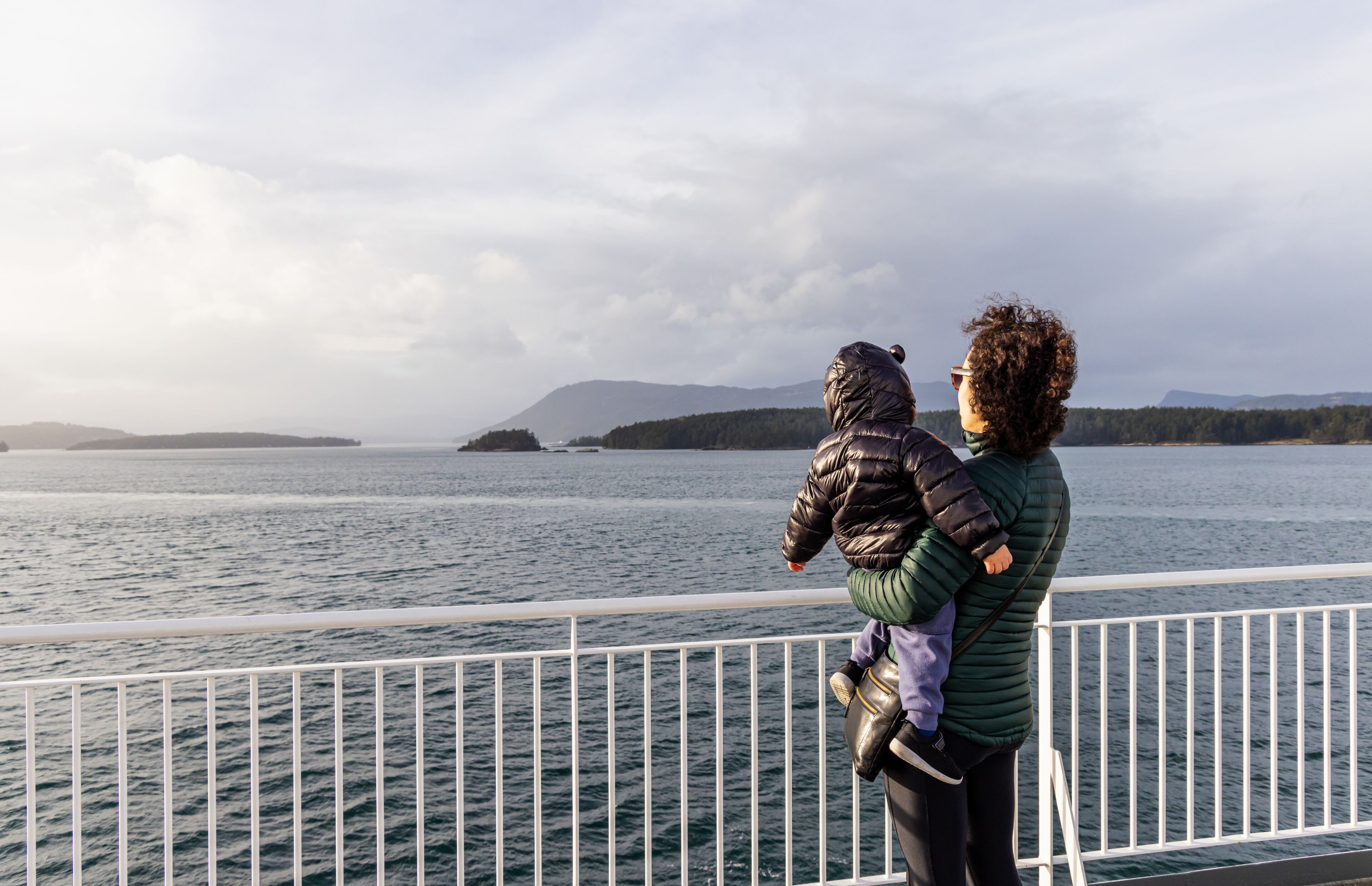 A person holding a child by a railing overlooking a body of water with distant landforms.