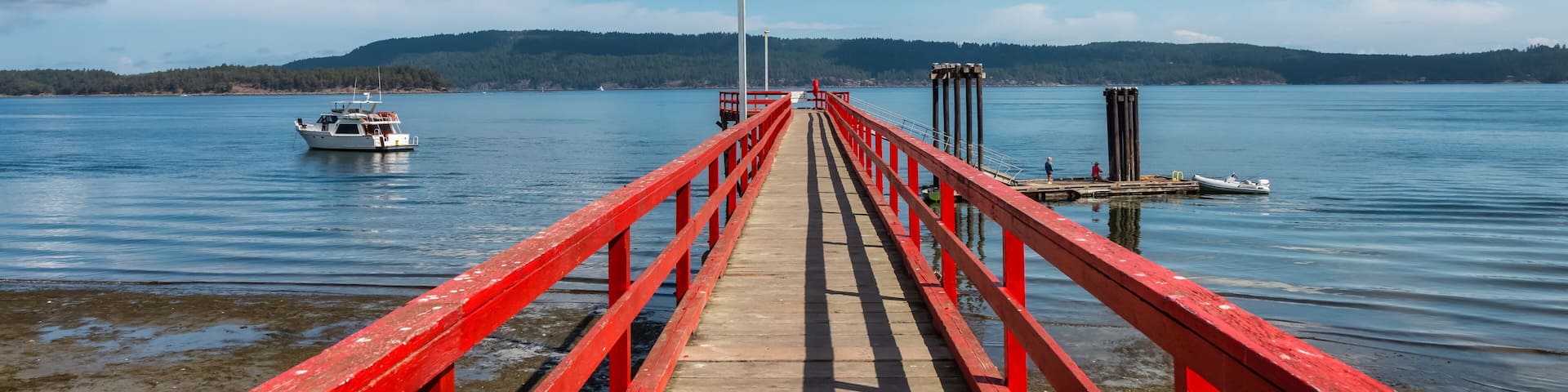 A wooden pier with red railings extending into a calm lake, surrounded by hills and a clear blue sky.
