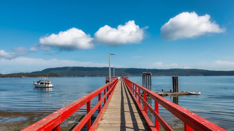 A wooden pier with red railings extending into a calm lake, surrounded by hills and a clear blue sky.