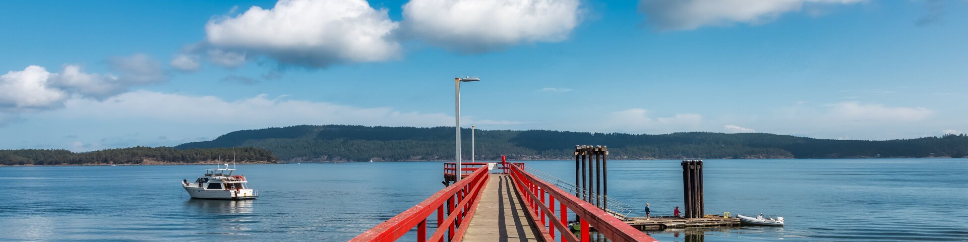 Um cais de madeira com guarda-corpos vermelhos estendendo-se sobre um lago calmo, rodeado por colinas e um céu azul límpido.