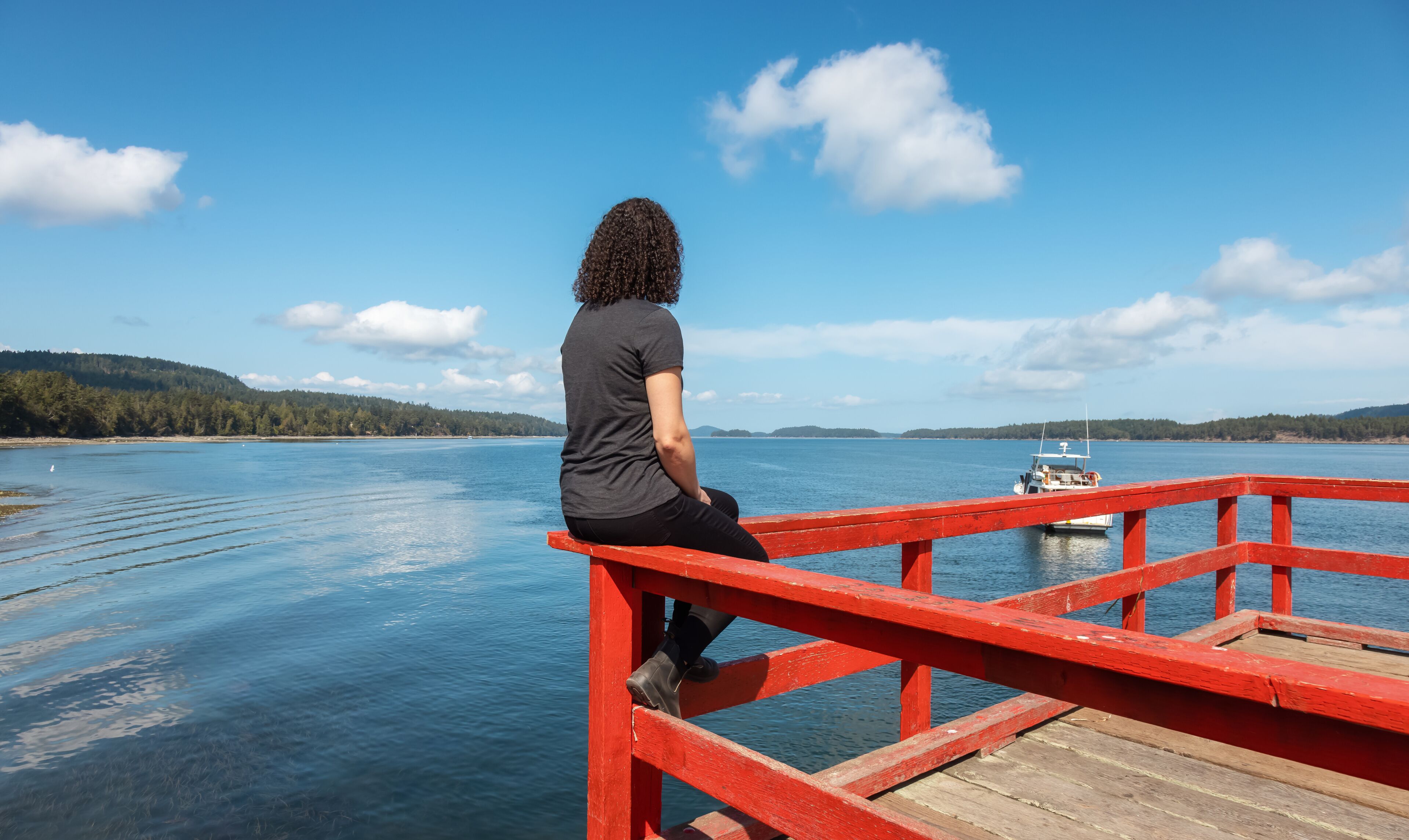 A person sitting on a red pier overlooking a calm lake with a boat in the distance and forested hills in the background.