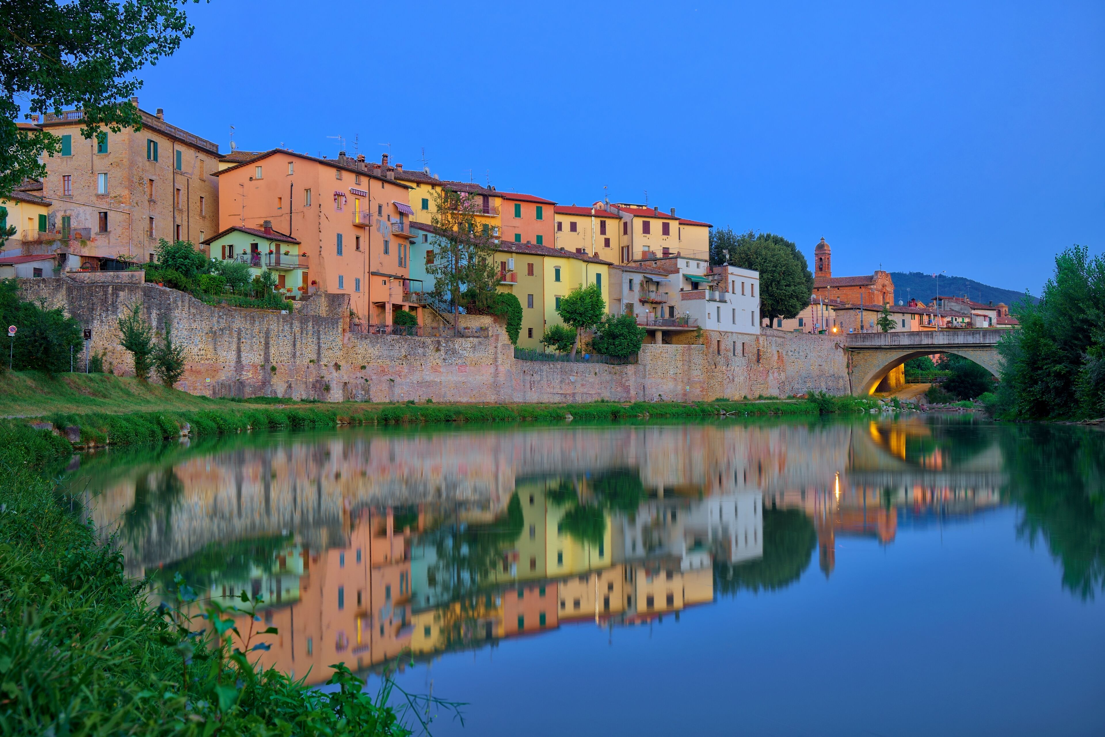 A calm river reflecting colorful buildings, a stone wall, and a bridge.