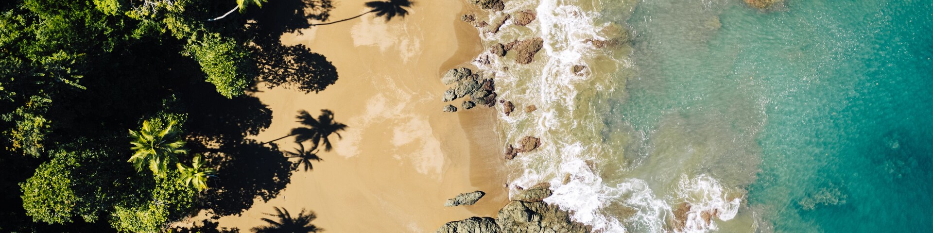 Aerial view of where the deep turquoise ocean meets the sandy shore, embraced by dark rocks and the lush green forest, Puerto Jimenez, Puntarenas Province, Costa Rica.