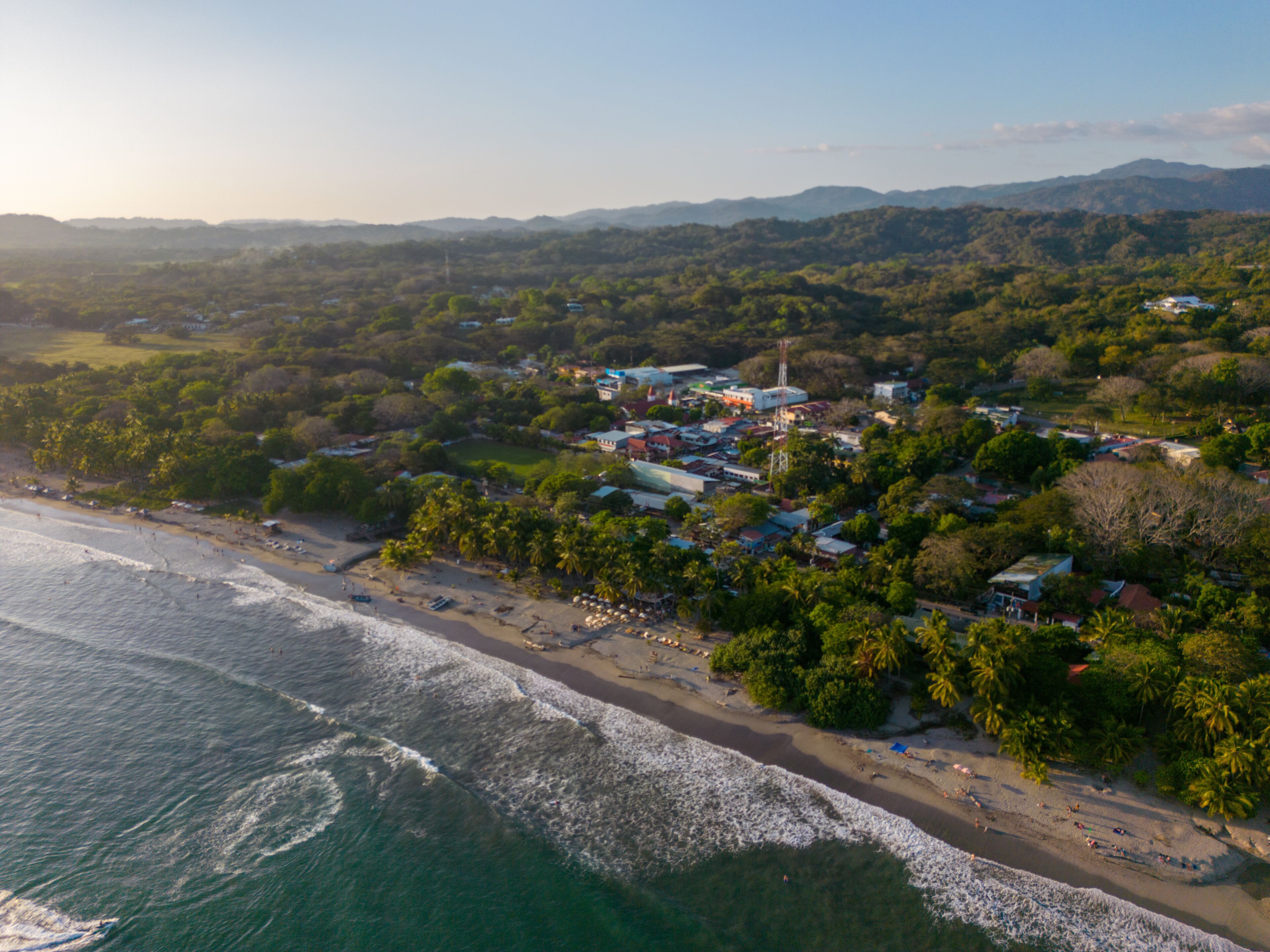 An aerial view of a coastal town with a beach, palm trees, and a mix of residential and commercial buildings.