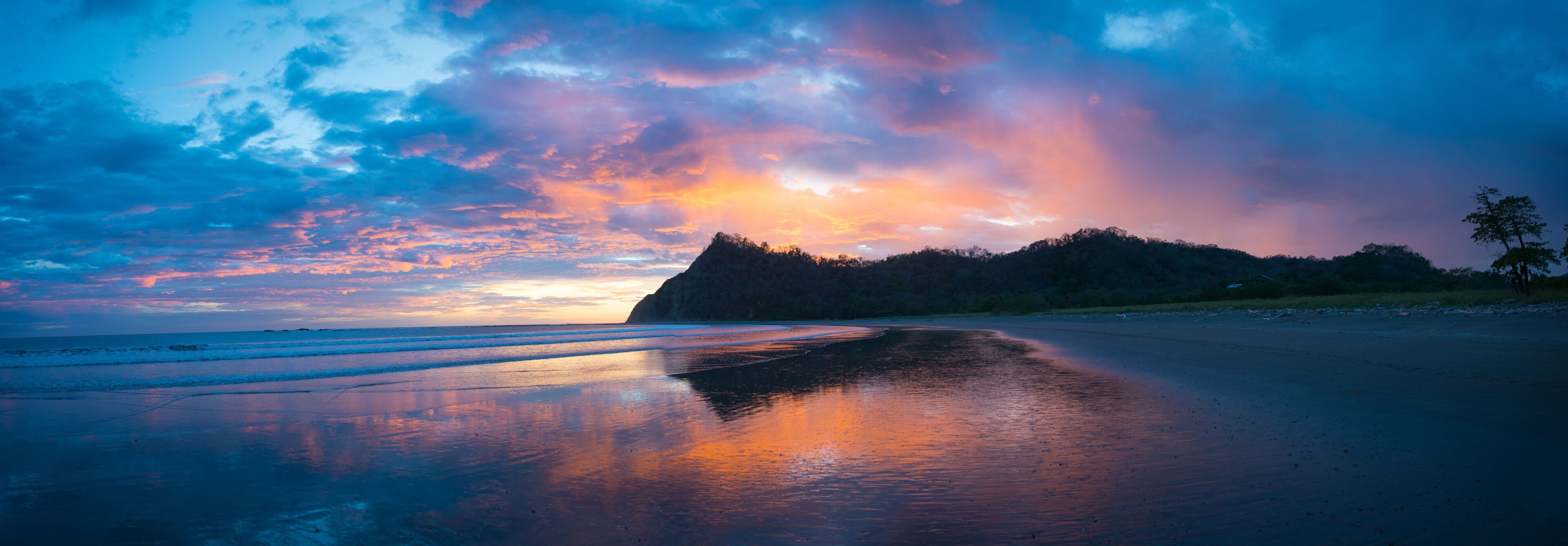 A beach with a reflective wet sand surface, a distant mountain, and a colorful sky.