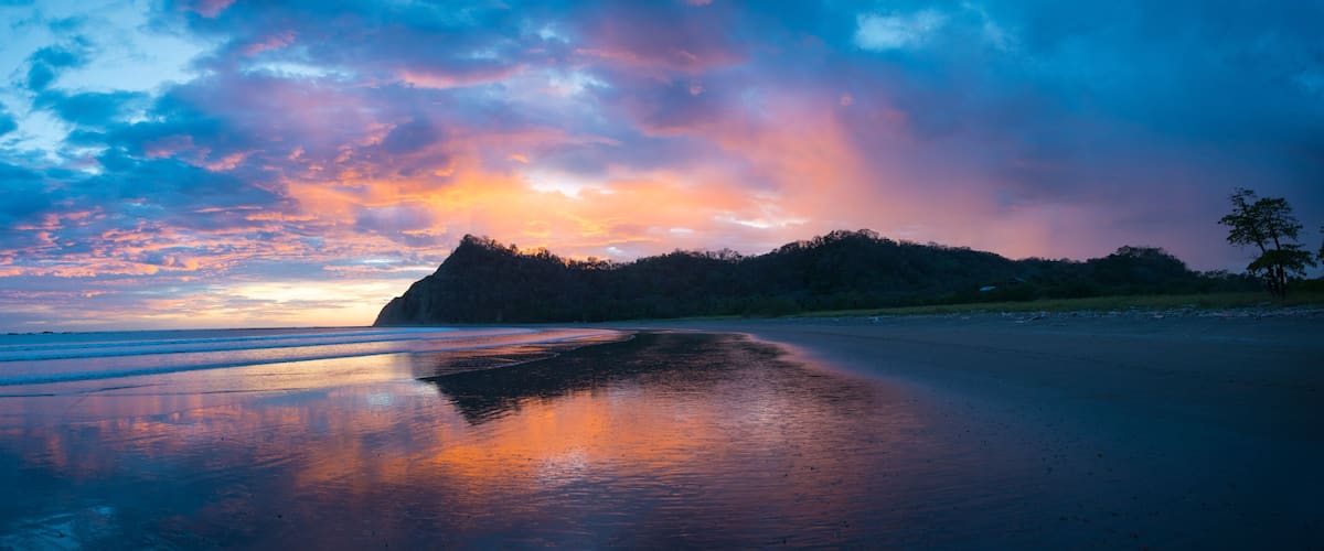 A beach with a reflective wet sand surface, a distant mountain, and a colorful sky.