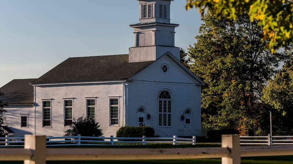 A white church with a steeple and a cross on top, surrounded by trees and a white fence.
