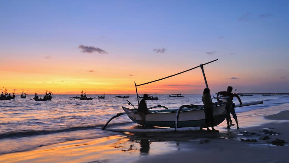 A sunset over a beach with several boats and people near the shore.