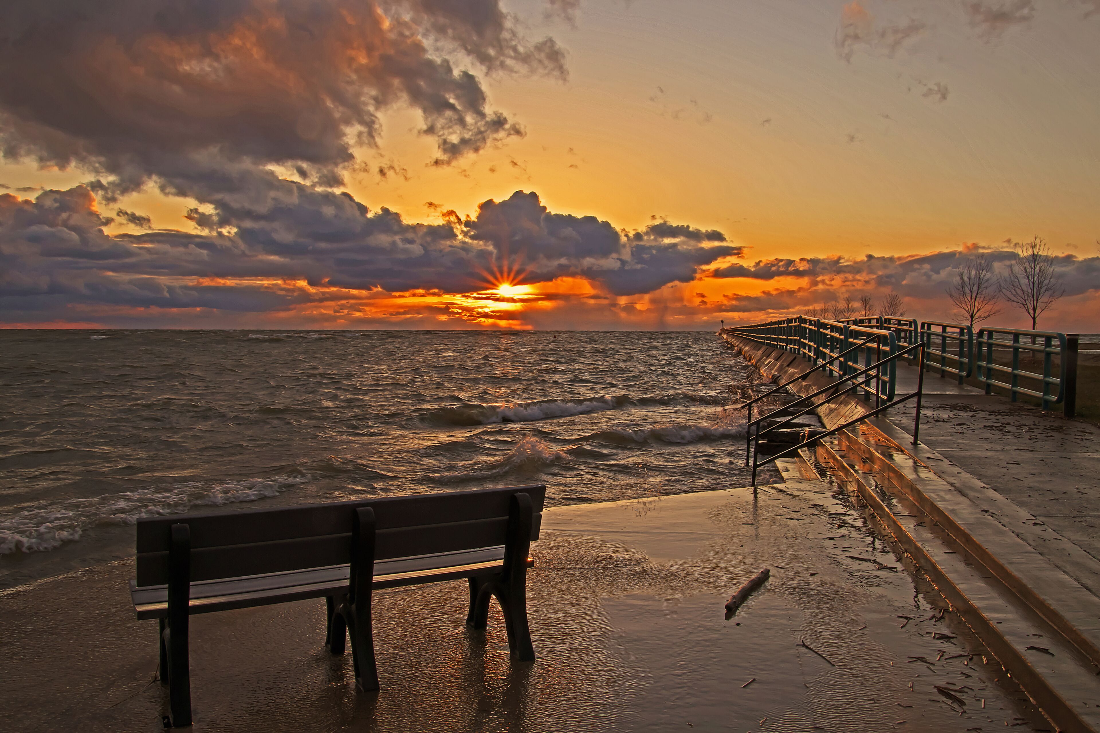 A sunset over the ocean with a bench and a boardwalk.