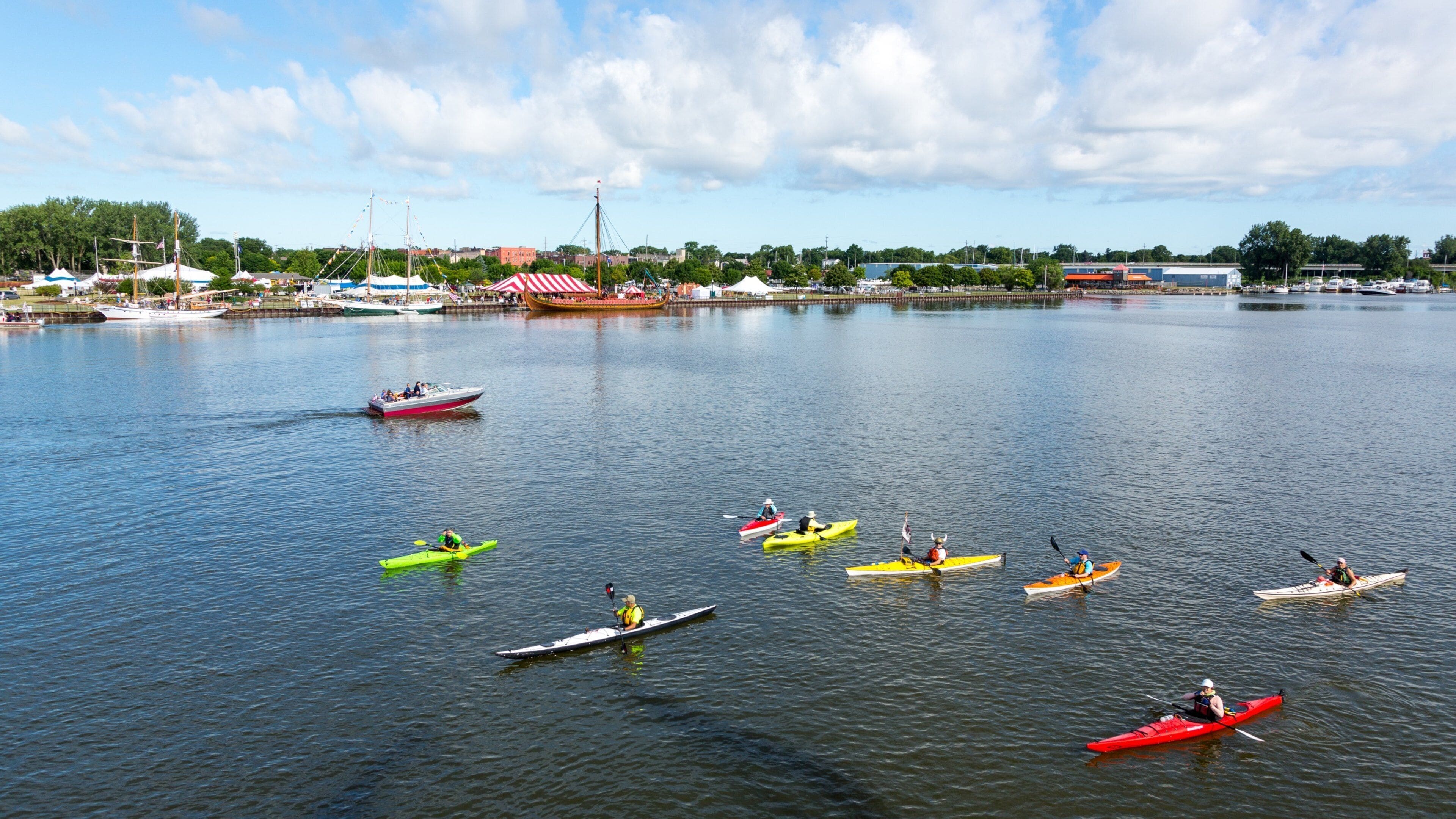 A calm lake with several kayaks and a speedboat. There are tents and buildings in the background.