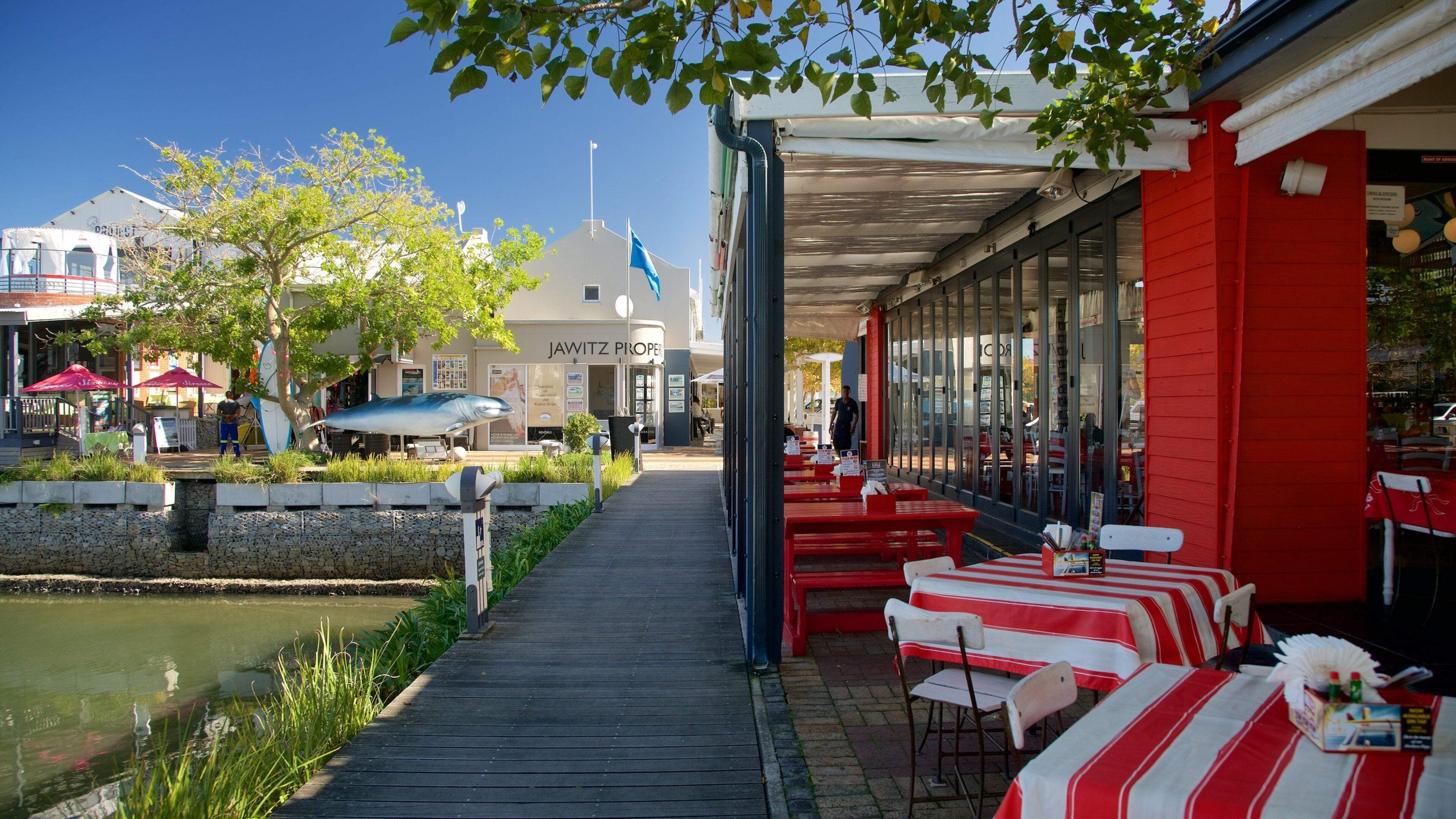 Um restaurante à beira do rio com mesas e cadeiras em listras vermelhas e brancas, um cais e uma grande escultura de tubarão.