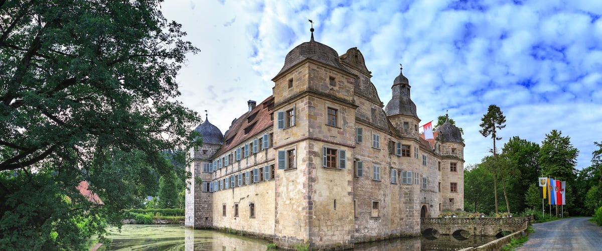 A historic stone castle with multiple towers, a moat, and flags.