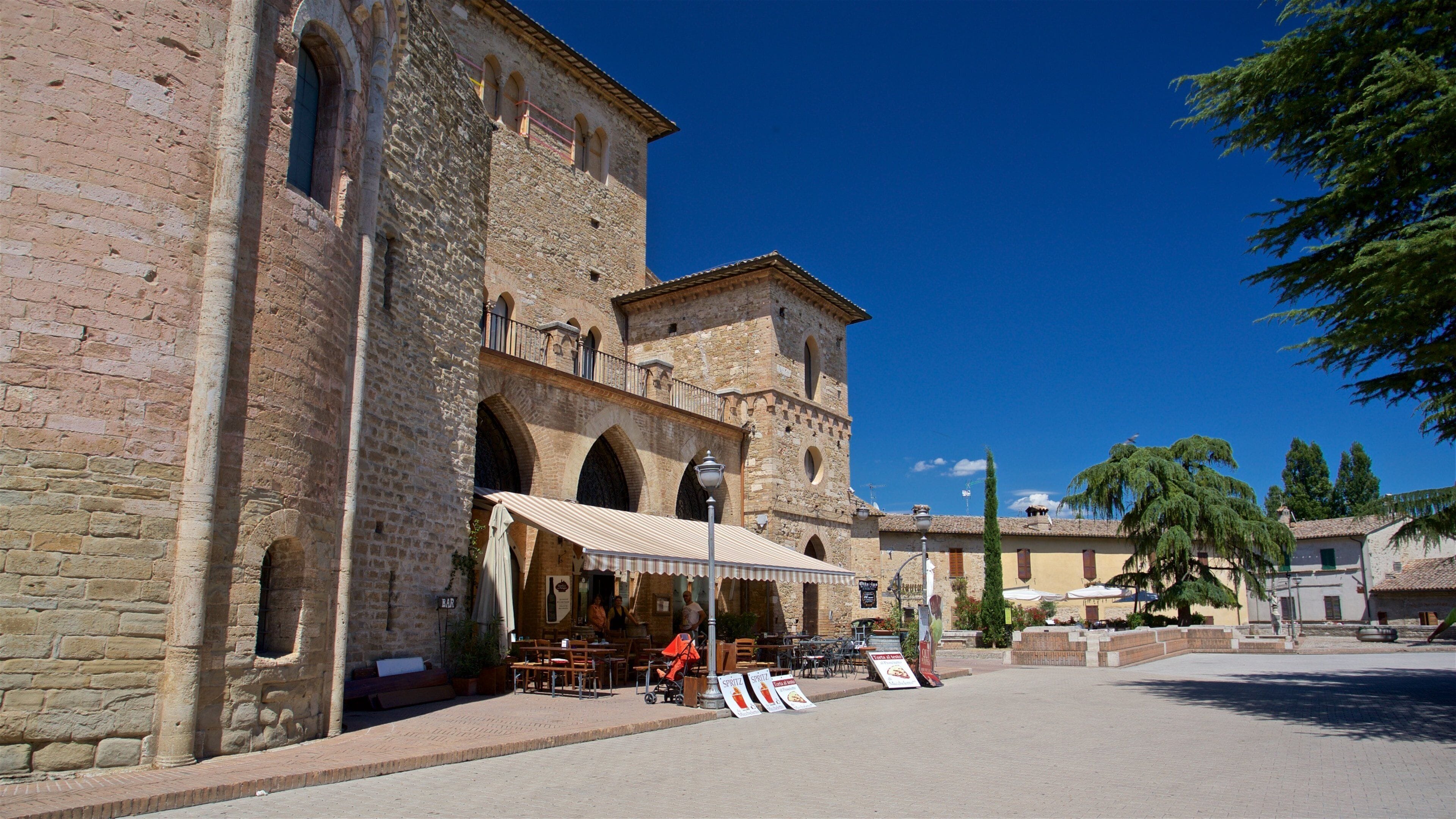 A historic stone building with arches and a tower, a café with outdoor seating, and a clear blue sky.