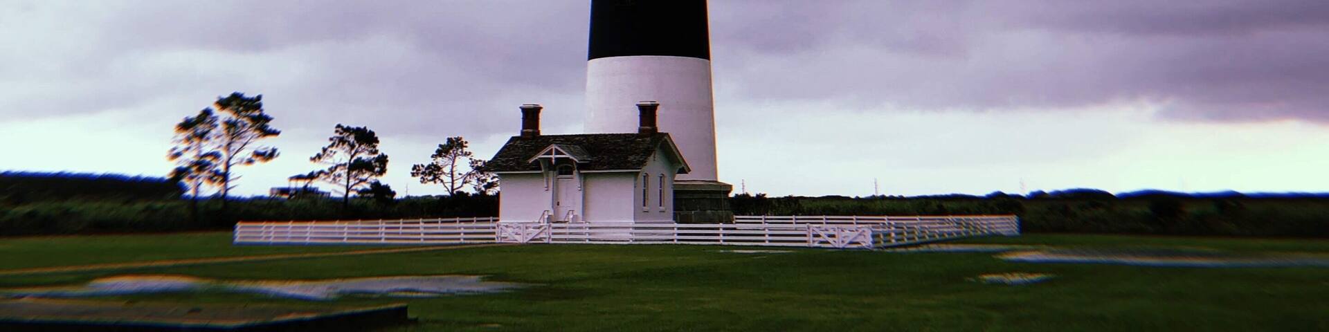 A tall, black and white striped lighthouse with a small building at its base, surrounded by a grassy field and trees under a cloudy sky.