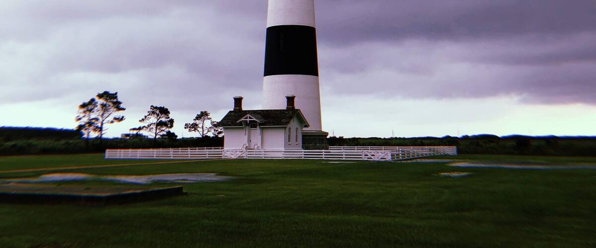 A tall, black and white striped lighthouse with a small building at its base, surrounded by a grassy field and trees under a cloudy sky.
