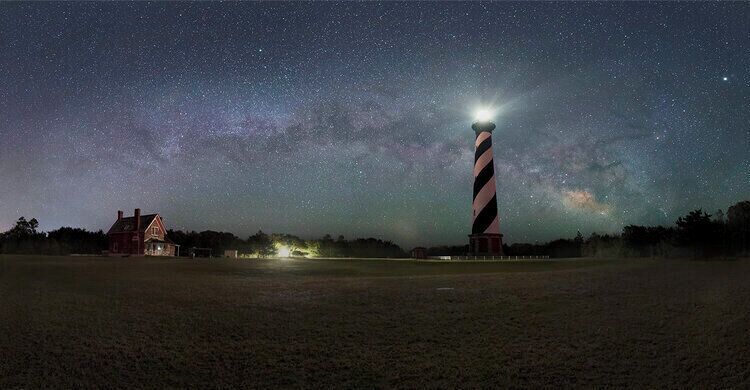 A lighthouse at night with a starry sky and a house in the foreground.