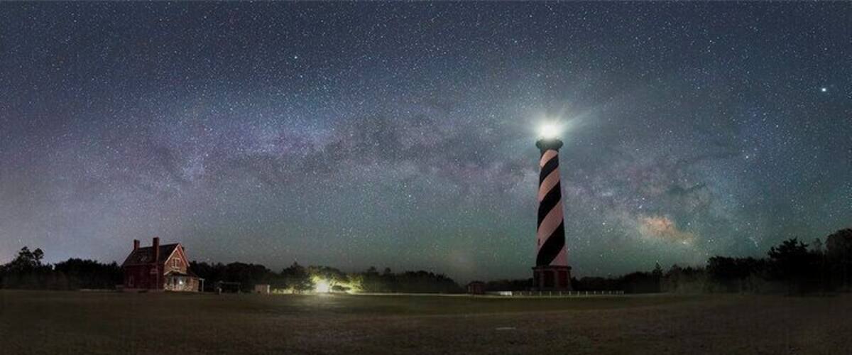 Un phare illuminé la nuit, avec un ciel étoilé et une maison au premier plan.