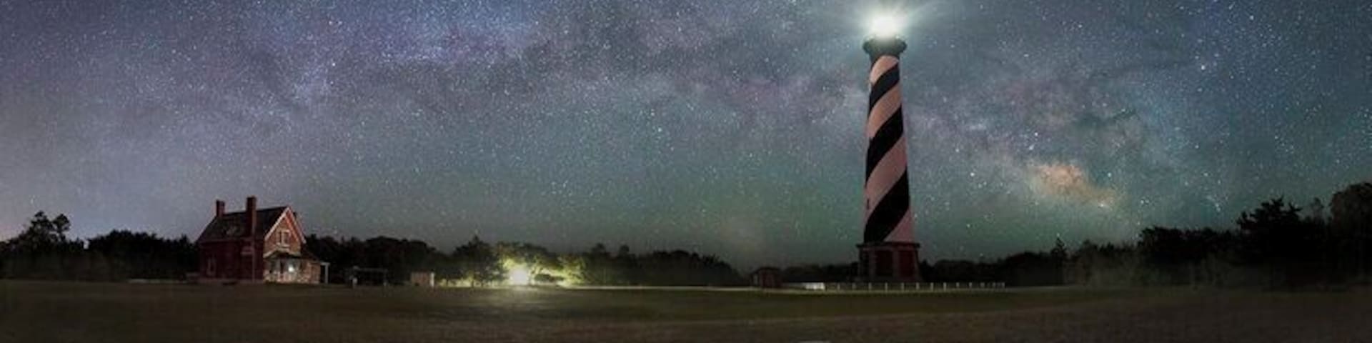 Un faro de noche bajo un cielo estrellado, con una casa en primer plano.