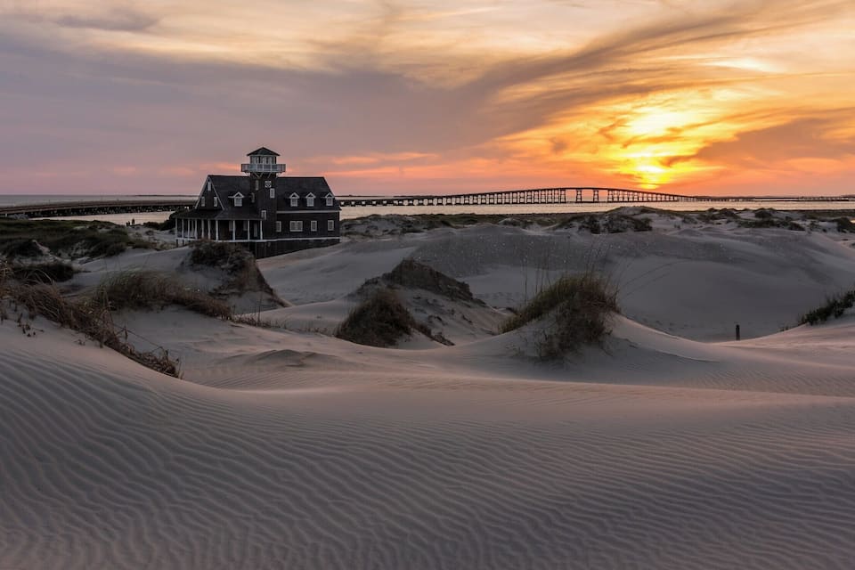 Uma casa com uma torre, dunas de areia e um pôr do sol sobre um píer.