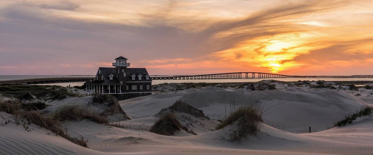 A house with a tower, sand dunes, and a sunset over a pier.
