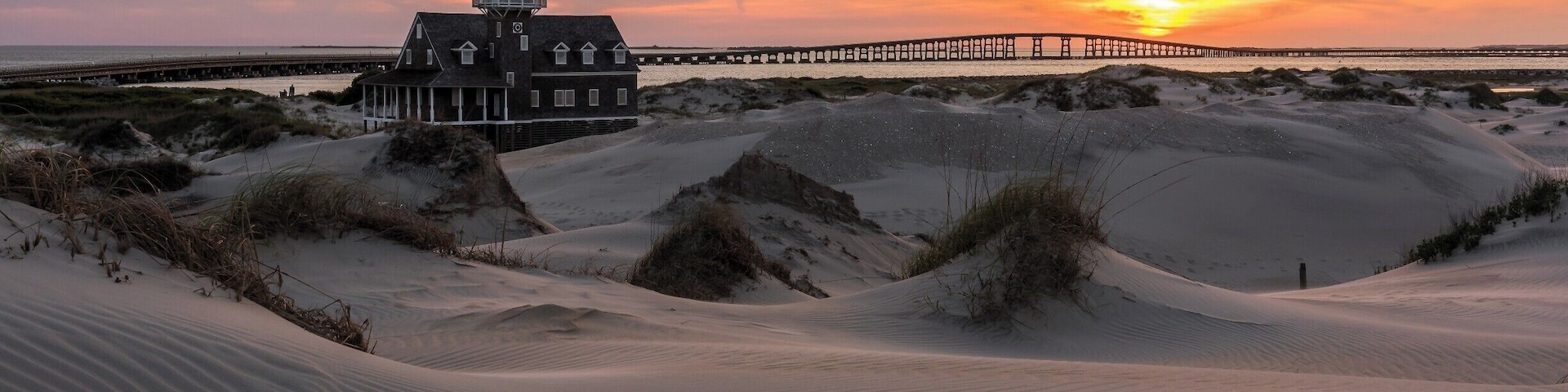 A house with a tower, sand dunes, and a sunset over a pier.