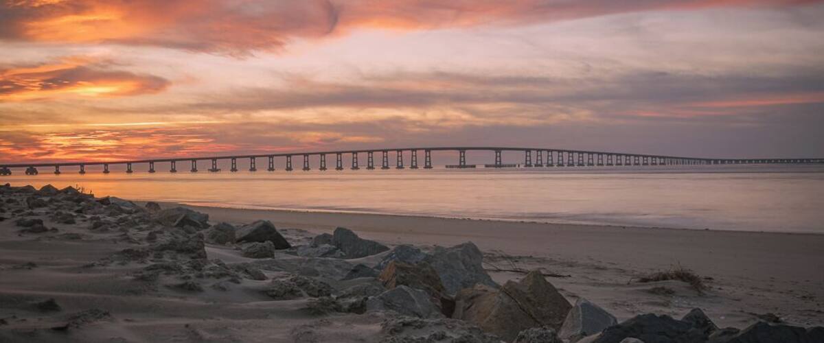 This image was captured at the Oregon Inlet on the NC Outerbanks in December of 2015. It features the iconic Bonner Bridge, one of the more famous bridges in North Carolina. This was an impromptu trip that I undertook hoping to catch a sight of Comet Catalina that was gracing our skies at the time. That trip turned out to be one of the best trips I have ever undertaken as I encountered perfect conditions for photography every single day over the 3 day period I was there.
This image is a blend of 2 images to bring out some detail in the foreground rocks. This composition appealed to me since the foreground rocks form a natural line to lead the viewer's eyes to the bridge and the clouds above. The clouds on this day were assuming various familiar shapes with the one captured here taking the shape of a bird in flight. 
This particular stretch of the shoreline is very peaceful to walk along. Since it's an inlet, the waves are extremely calm unless it's windy. There are several sand dunes to the left of this image that ultimately would lead you to the proper coast and the ocean. 
Update, 2020: This bridge is being dismantled and replaced with a newer bridge. Bonner Bridge has a lot of history tied to it and while it's sad to see it go, I feel fortunate to have captured part of this history personally.
Thank you for stopping by and for the support.
#sunset #nc #usa #bridge #obx #outerbanks #colorful #goldenhour #roadtrip #weekend #beach #beaches #bestof5 #nationalpark #bridges #lovemytown 
#history #BeachBound