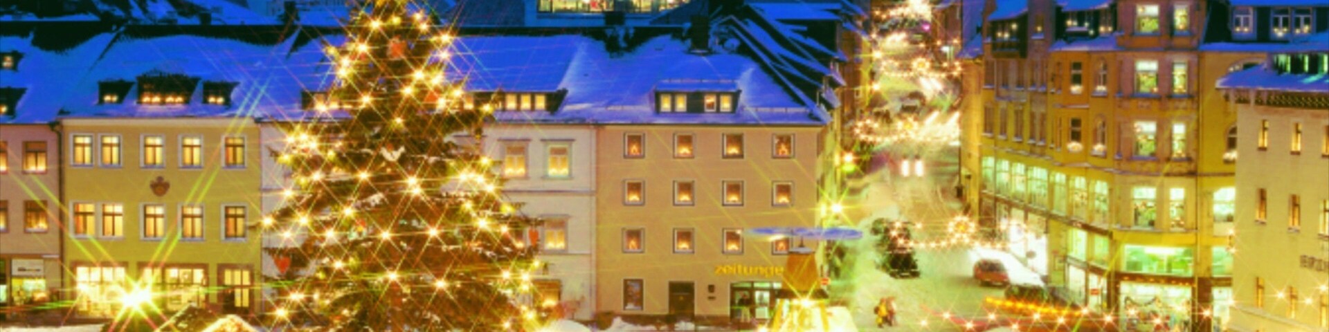 A snow-covered town square with a large Christmas tree and illuminated buildings.