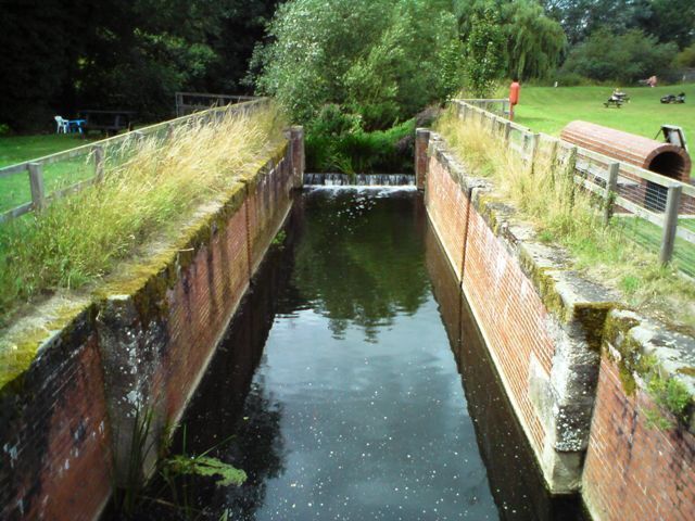 A narrow canal with brick walls, a grassy embankment, and a small waterfall.