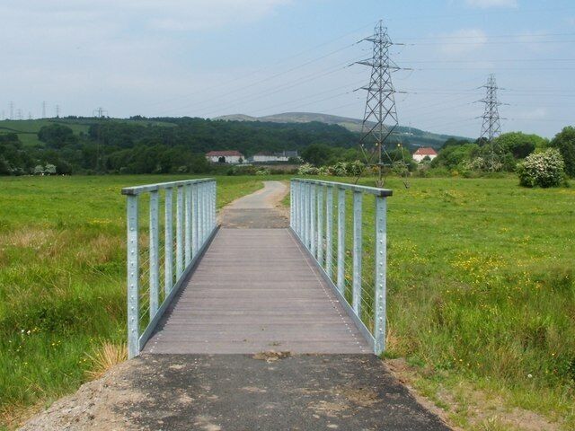 Uma ponte de madeira com guarda-corpos de metal numa paisagem campestre.