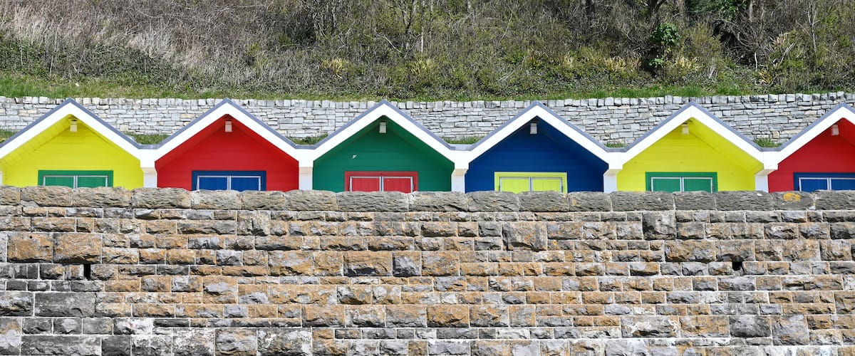A row of colorful beach huts with stone walls in the background.