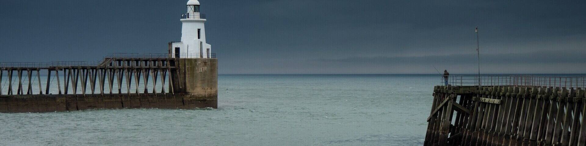 A white lighthouse on a pier extending into calm waters under a cloudy sky.