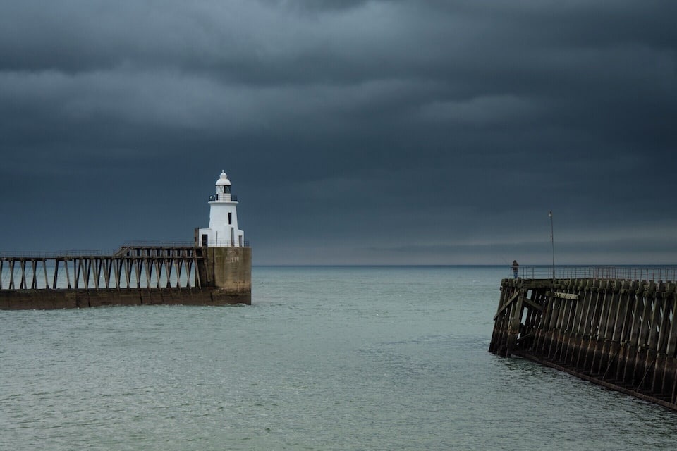 A white lighthouse on a pier extending into calm waters under a cloudy sky.