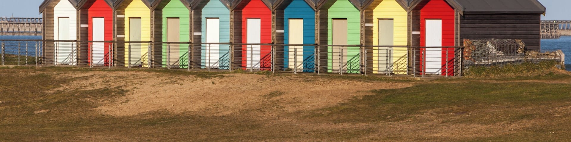 A row of colorful beach huts with a metal fence in front, set against a clear sky and a body of water in the background.