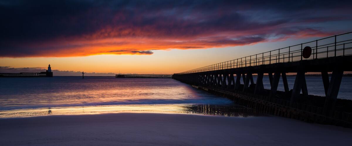 A pier extending into a calm sea at sunset with a dramatic sky.