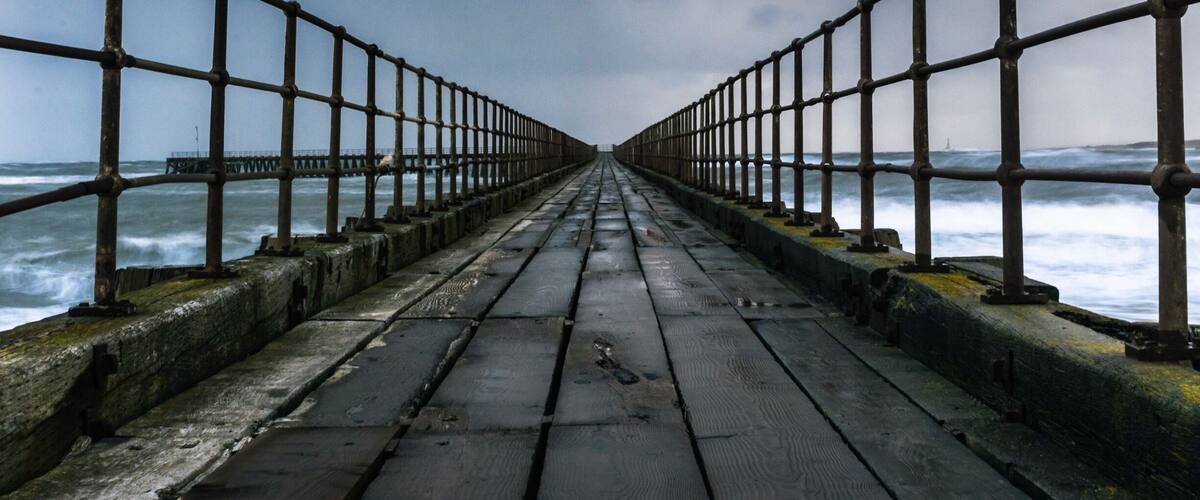 Une jetée en bois, dotée de rampes métalliques, s’avance dans une mer agitée.