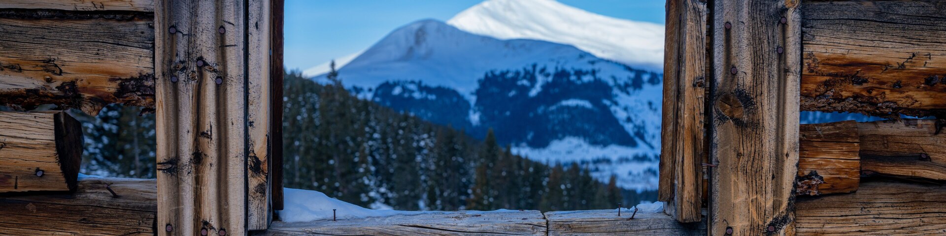 A wooden cabin with a window framed by logs, overlooking a snow-covered mountain range.