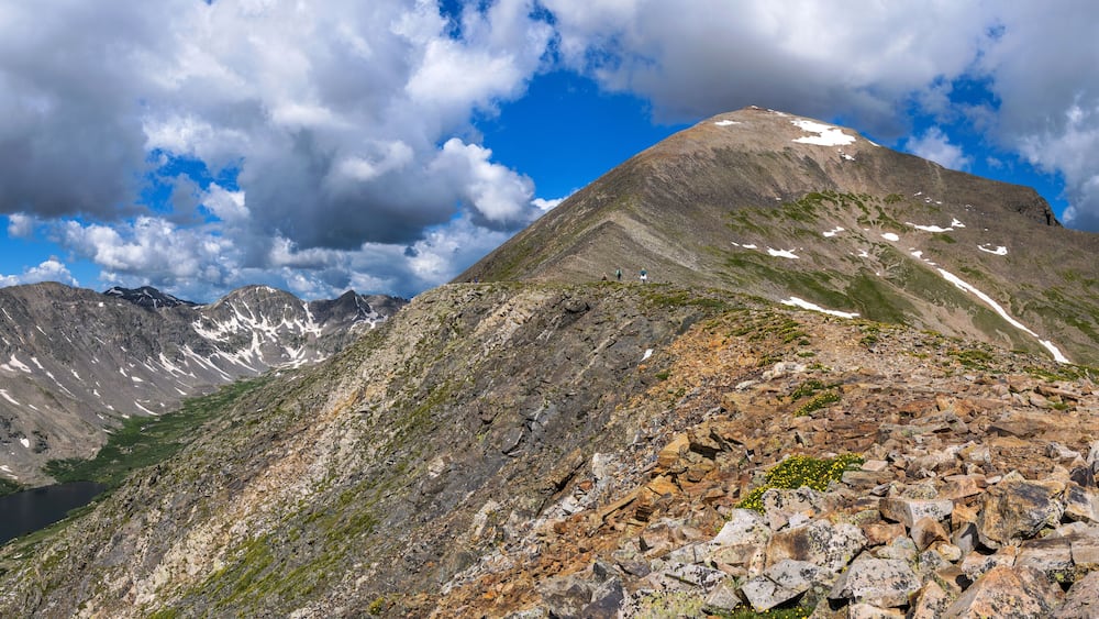 A mountain with a rocky foreground, a lake, and snow patches.