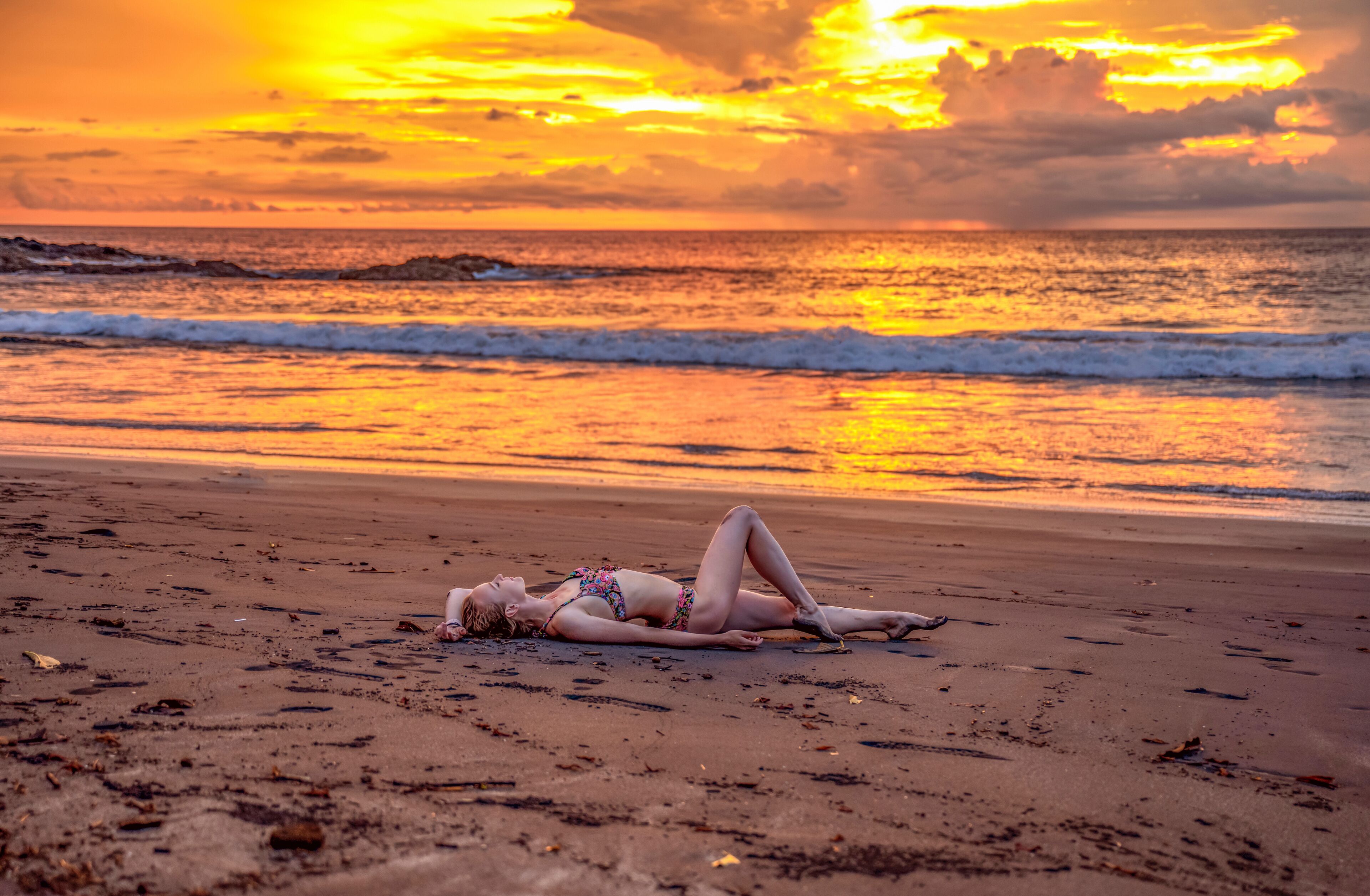 A beach with a sunset, waves, and a person lying on the sand.