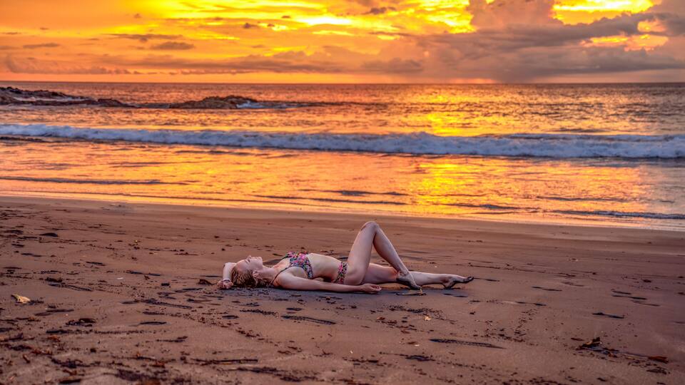 A beach with a sunset, waves, and a person lying on the sand.