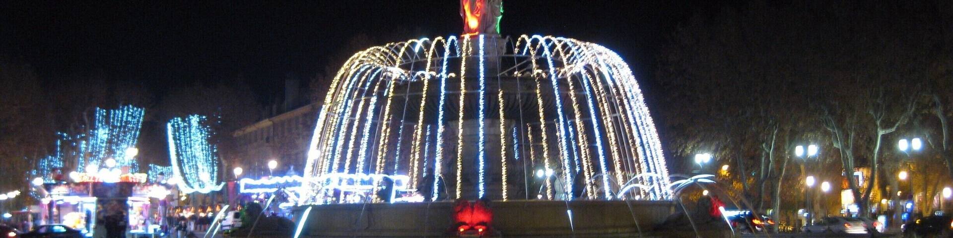 A brightly lit fountain with a statue in the center, surrounded by illuminated trees and buildings.