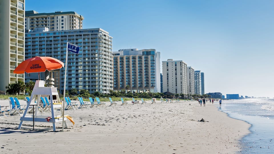 A beach with a lifeguard stand, beach chairs, and tall buildings in the background.