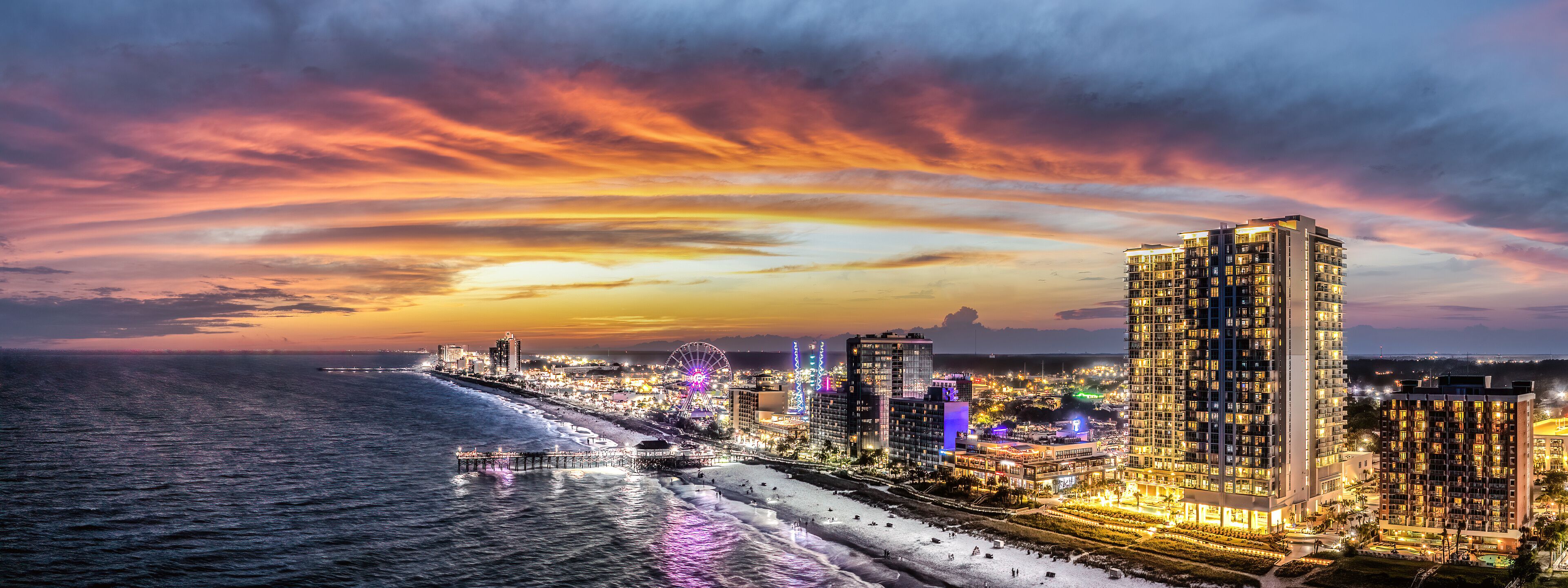 A coastal cityscape at sunset with illuminated buildings and a pier extending into the ocean.