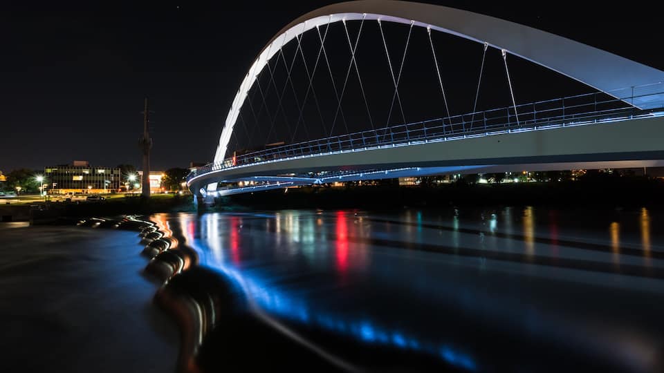 A modern bridge with a curved arch, illuminated lights, and reflections on the water.